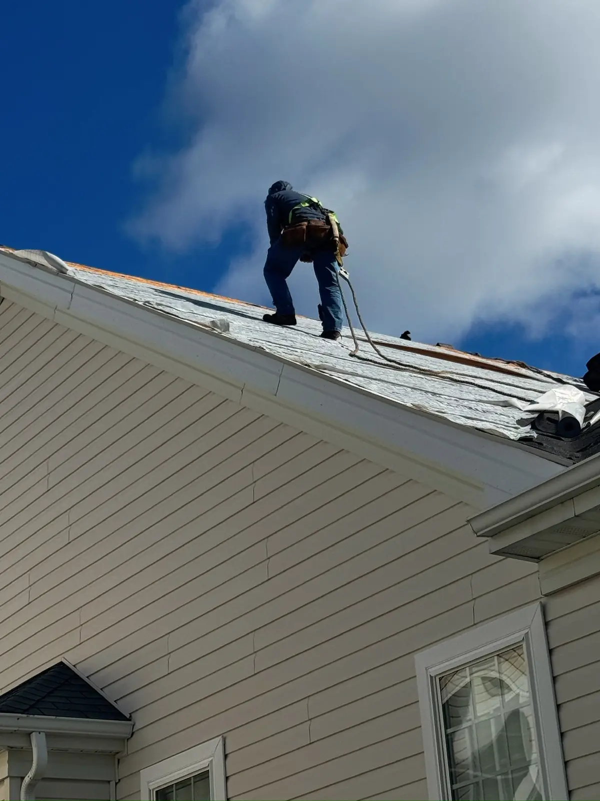 Roofer in a safety harness working near the ridge of a home under a blue sky