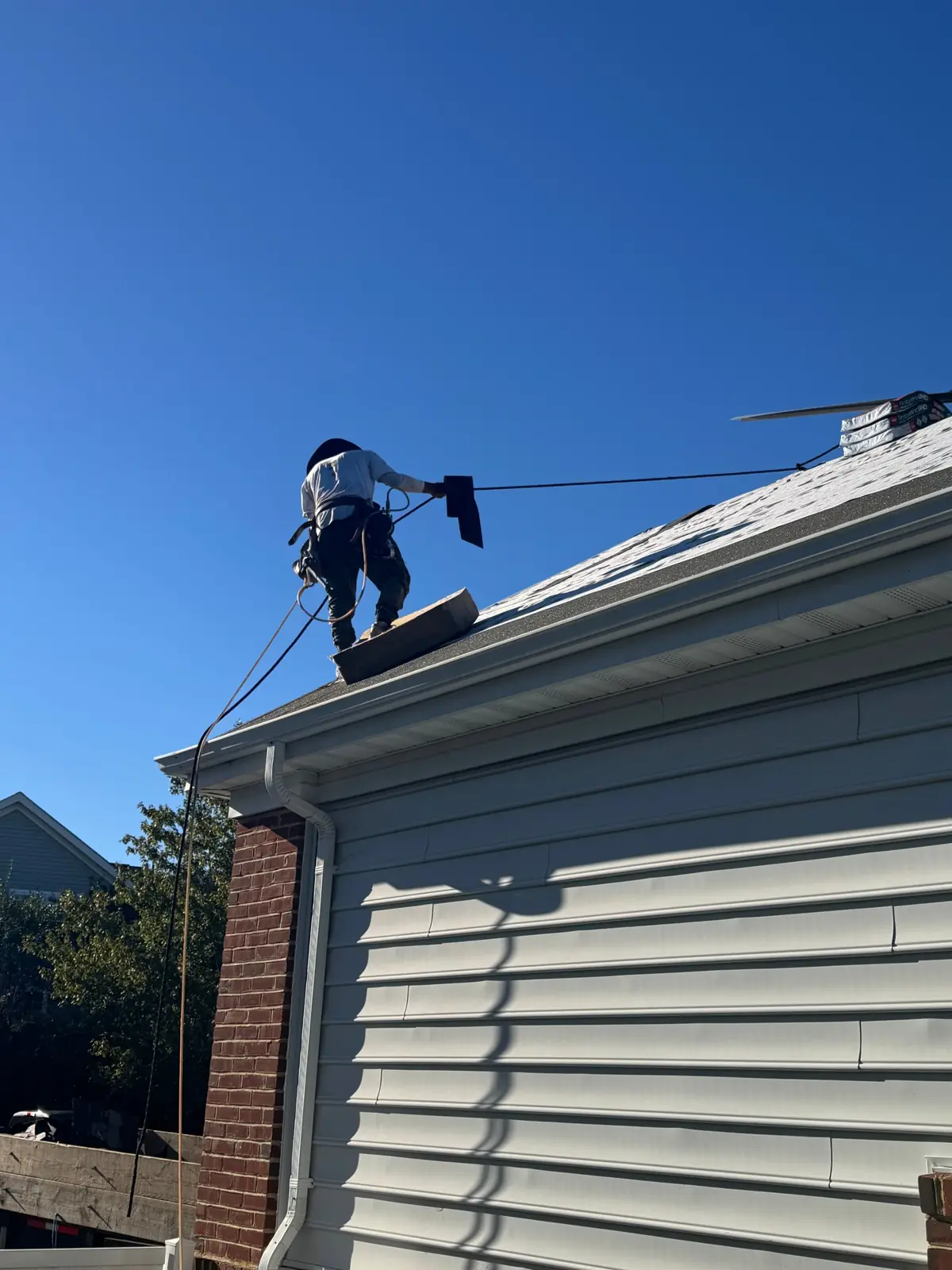 Roofer in a safety harness carrying a shingle bundle along a roof edge above white gutters